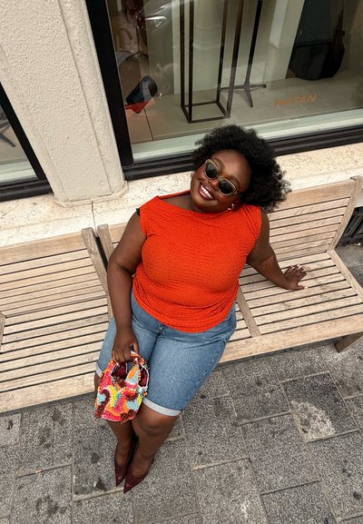Mujer sonriente con gafas de sol, vestida con una blusa naranja y shorts de mezclilla, sentada en un banco de madera con una bolsa de cuentas colorida en la acera de la ciudad.