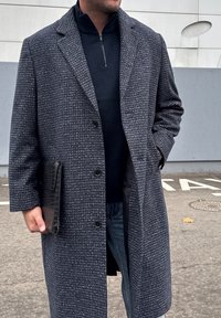 Manteau gris texturé avec un col cranté, doté de deux boutons, d'une poche unique, et d'un pull noir à fermeture éclair en dessous.
