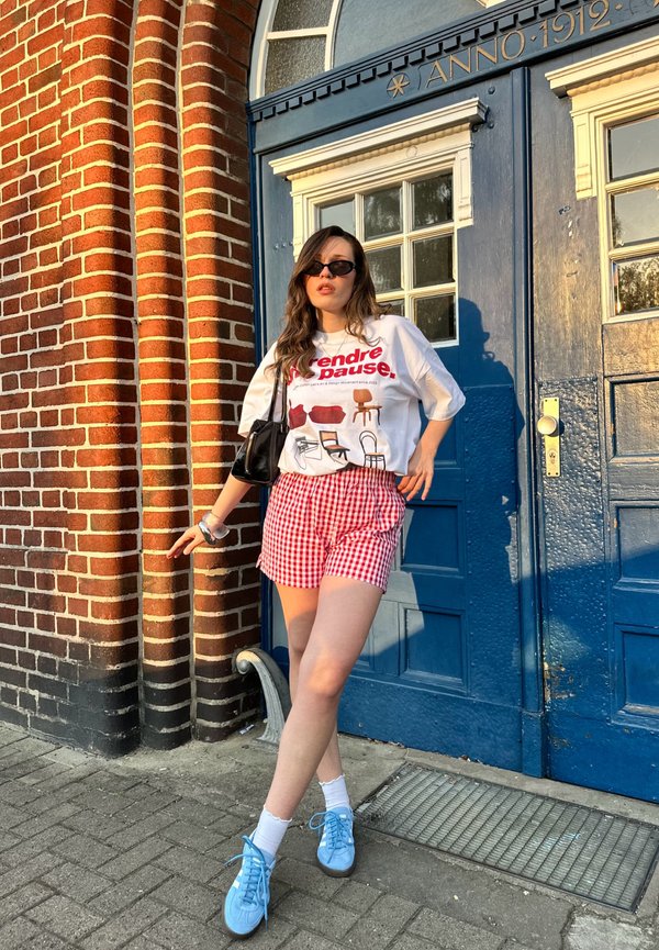 Camiseta blanca con diseño gráfico, pantalones cortos de cuadros rojos, zapatillas azul claro y un bolso negro, frente a una puerta azul y una pared de ladrillo.