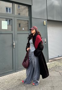 Dlhý čierny kabát s červenými akcentmi, sivý top, sivé široké nohavice, bordová taška a červené topánky. Hnedá čiapka a tmavé vlasy, stojaci pri šedej dverách.