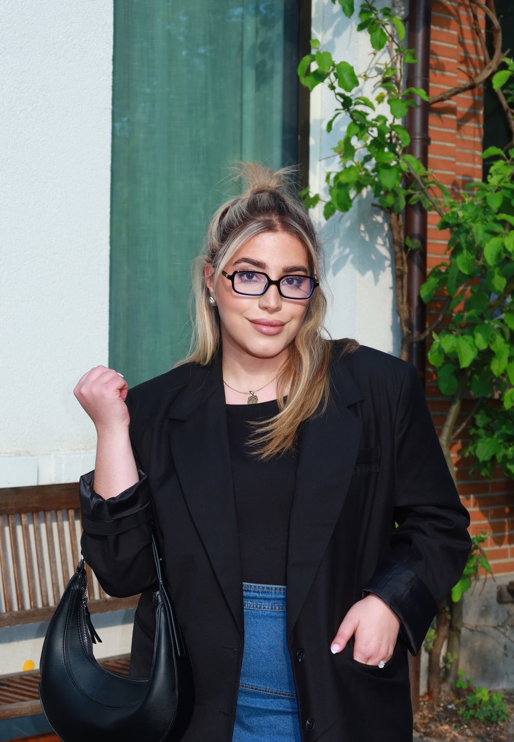 Czarny żakiet na czarnej bluzce, niebieska spódnica dżinsowa i mała czarna torebka. Długie proste blond włosy, nosi okulary i naszyjnik z zawieszką.