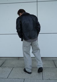 Zwarte leren jas, grijze sweatshirt en lichtgrijze brede jeans. Staand tegen een witte muur, met een eenvoudig ontwerp en gedempte kleuren.