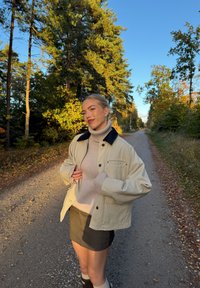 Beżowa bawełniana kurtka z granatowym kołnierzem, jasnoszary sweter z golfem, ciemna spódnica i skarpety do kolan, umieszczone na żwirowej ścieżce otoczonej drzewami.