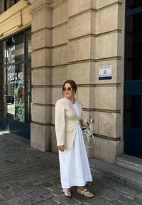 Μπεζ σακάκι πάνω από ένα άσπρο μάξι φόρεμα, διαφανή παπούτσια, κρατώντας ένα ποτό. Γυαλιά ηλίου και μια ελαφριά τσάντα. Φόντο από πέτρινο τοίχο και παράθυρο.