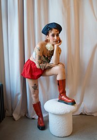 Punaiset shortsit, raidallinen crop-top, ja pitkät punaiset nahkasaappaat vetoketjulla, asusteena sininen baretti ja valkoinen kukkakoriste. Valkoinen pörröinen jakkara.