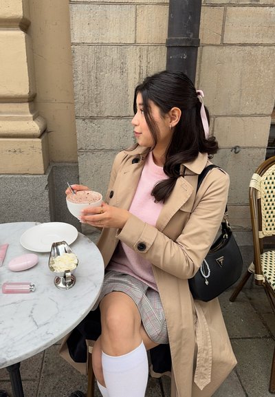 Abrigo beige sobre un suéter rosa, sentado en una mesa de mármol. Sosteniendo un cuenco blanco de postre con una cuchara. Falda de cuadros y calcetas hasta la rodilla.