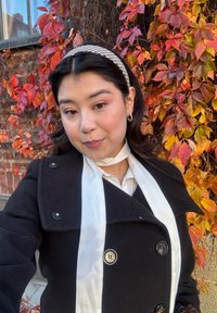 Zwarte jas met grote knopen en een witte sjaal. De achtergrond toont levendige rode en oranje bladeren op een muur. Glad textuur, warme tonen.