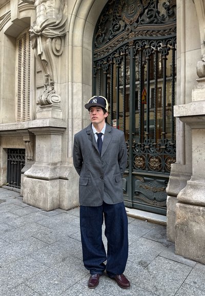 Blazer de tela gris con dos botones, combinado con pantalones de mezclilla azul sueltos. Gorra negra de los New York Yankees y zapatos color burdeos. Fondo de edificio de piedra.