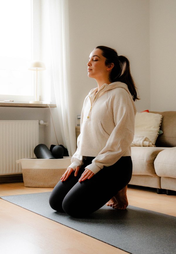 Gaiši beidžs, īsais kapuce ar priekšējo kabatu un zelta rāvējslēdzēju; melnas legingas; ceļos uz pelēka jogas paklāja gaišā iekštelpu vidē.