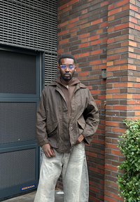 Braune Zip-Jacke mit zwei Fronttaschen über einem braunen Hoodie, kombiniert mit hellgrauen, weit geschnittenen Jeans. Auffällige, blau getönte Brille.