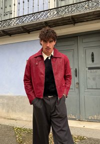 Νέος άνδρας με κατσαρά μαλλιά που φοράει κόκκινο μπουφάν, μαύρο πουλόβερ, λευκό πουκάμισο και φαρδιά σκούρα παντελόνια στέκεται μπροστά από γκρι διπλές πόρτες.