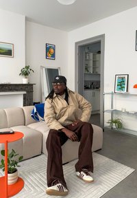 Bēša jaka, brūnas plašas bikses un baltas sporta kurpes ar tumši sarkaniem svītrām. Pelēks dīvāns, oranža sānu galdiņa, zaļa augs un abstrakta māksla.