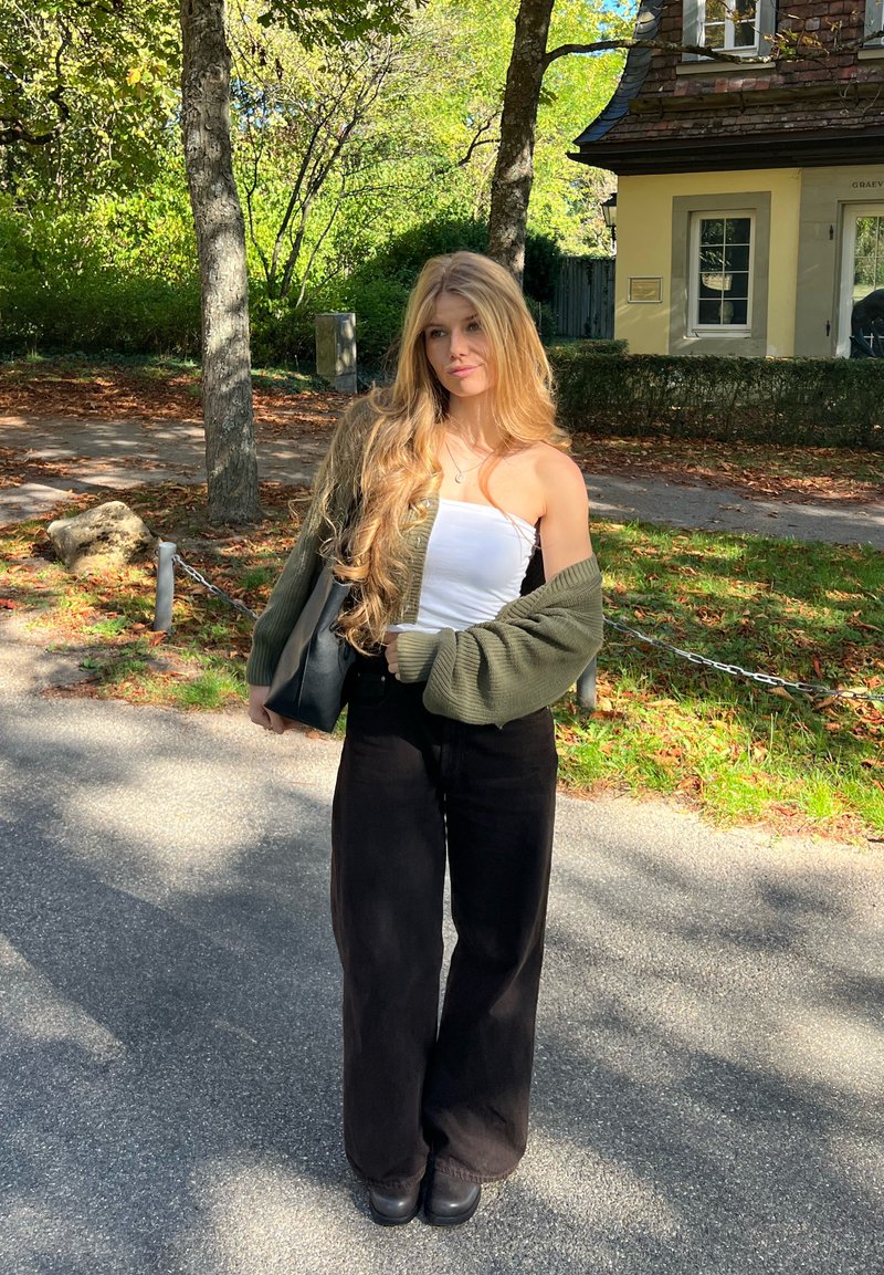 Jeune femme aux longs cheveux blonds portant un haut bandeau blanc, un cardigan vert et un pantalon noir large, debout sur un sentier ensoleillé près des arbres et d’un bâtiment.