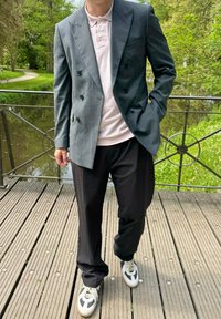 Blazer doppiopetto grigio carbone, polo rosa chiaro, pantaloni neri larghi e scarpe da ginnastica bianche con dettagli neri, in piedi su un ponte di legno.