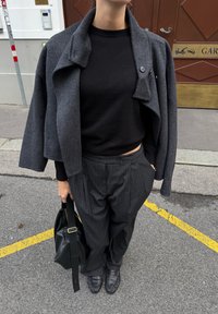 Černý fitted svetr spárovaný se šedou texturovanou zkrácenou bundou a pruhovanými širokými kalhotami. Doplňky zahrnují tmavou tašku a mokasíny.