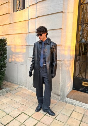 Hombre con abrigo de cuero negro, guantes, gafas de sol, camisa azul, corbata y pantalones de pierna ancha de pie sobre un pavimento de baldosas cerca de un edificio de piedra al atardecer.