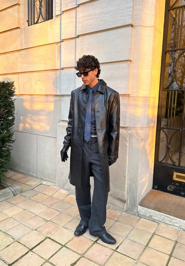 Mand iført sort læderjakke, handsker, solbriller, blå skjorte, slips og bukser med vide ben, stående på flisebelagt fortov nær stenhøj bygning ved solnedgang.