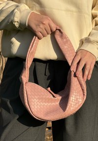 Bolsa de couro rosa trançado com uma forma curva, uma alça grossa e fecho de zíper. Superfície texturizada com um detalhe de padrão sutil.