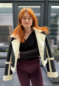 Jeune femme aux longs cheveux roux, portant un pull noir et une veste en peau lainée posée sur les épaules, debout à l'intérieur près de grandes fenêtres.