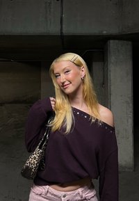 Ung kvinna med blont hår iklädd en mörk avskuren tröja och ljusrosa byxor, som håller i en väska med leopardmönster i ett svagt upplyst betongutrymme.
