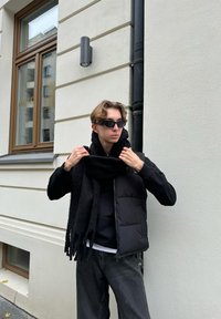 Zwarte gewatteerde vest over een zwarte hoodie, met een zwarte franjesjaal. Donkergrijze jeans en zwarte zonnebril. Eenvoudige stadsachtergrond.