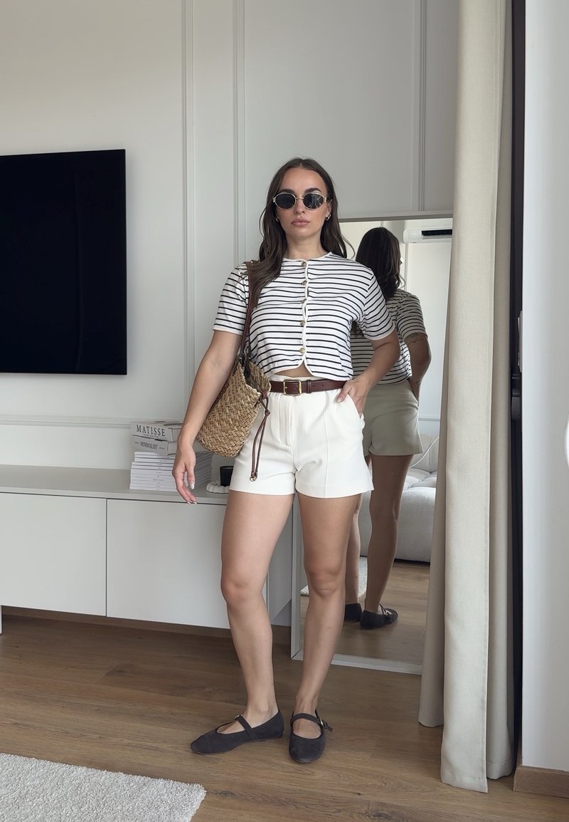 Femme portant des lunettes de soleil rondes, une chemise blanche à rayures boutonnée, un short blanc, des chaussures plates noires, tenant un sac tissé et debout dans une pièce moderne avec un miroir.