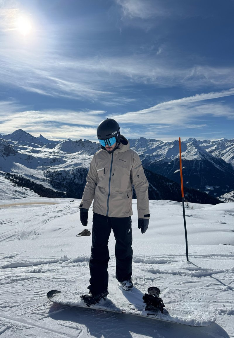 Homme portant un casque et des lunettes de protection, debout sur un snowboard sur une montagne enneigée, avec le soleil et un ciel bleu au-dessus.