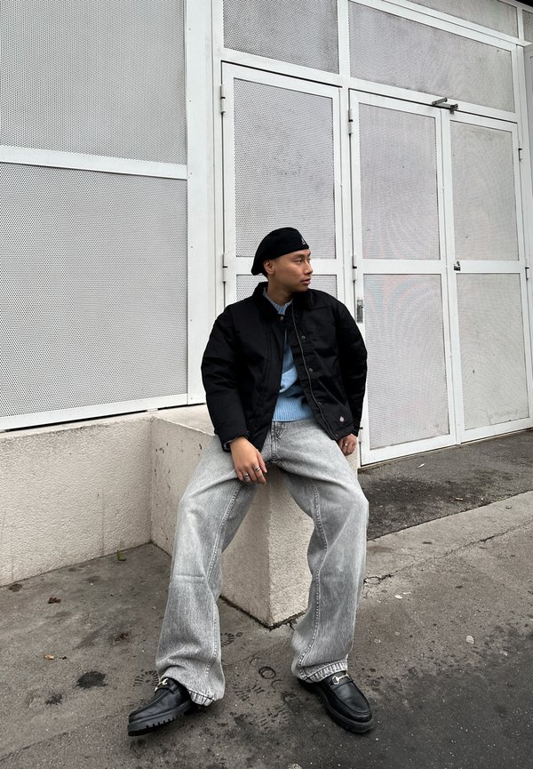 Giacca imbottita nera sopra un maglione azzurro chiaro, abbinata a jeans in denim grigio larghi e mocassini neri. L'ambientazione presenta uno sfondo bianco texturizzato.