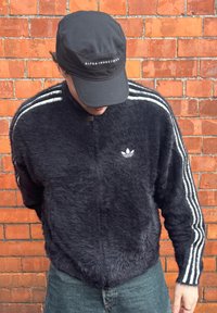 Fekete bolyhos dzseki fehér csíkokkal az ujjain és az Adidas logóval. A személy fekete sapkát visel, amelyen az "ALPHA INDUSTRIES" felirat olvasható.