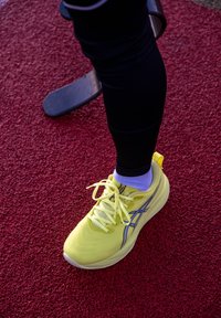 Helles gelbes Sportschuh mit reflektierenden silbernen Akzenten, strukturierter Oberseite und flachen Schnürsenkeln, getragen mit schwarzen Leggings auf einer roten Laufbahn.