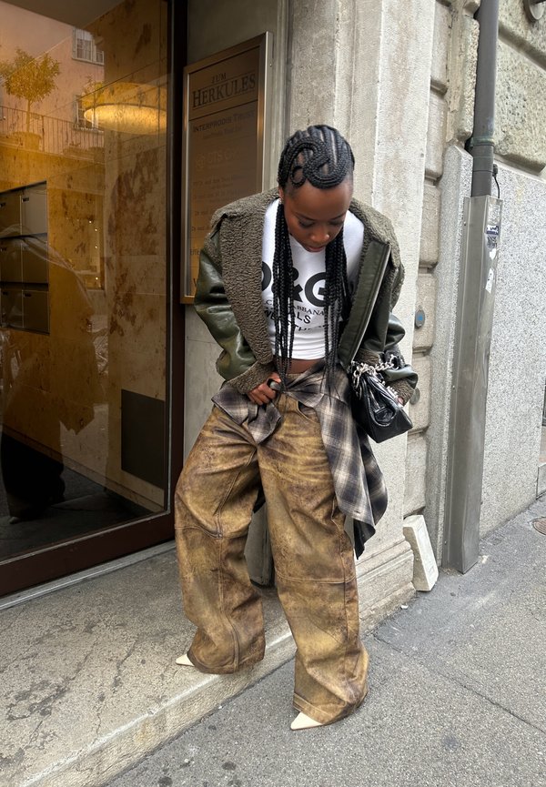 Chaqueta bomber de piel sintética color verde oliva forrada, top corto blanco, pantalones de textura marrón oversize, camisa de cuadros negra atada a la cintura, zapatos beige de punta.