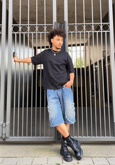 Camiseta de algodón negra, pantalones cortos de mezclilla azul claro con acentos blancos, y botas de combate negras, apoyándose contra una puerta de metal.