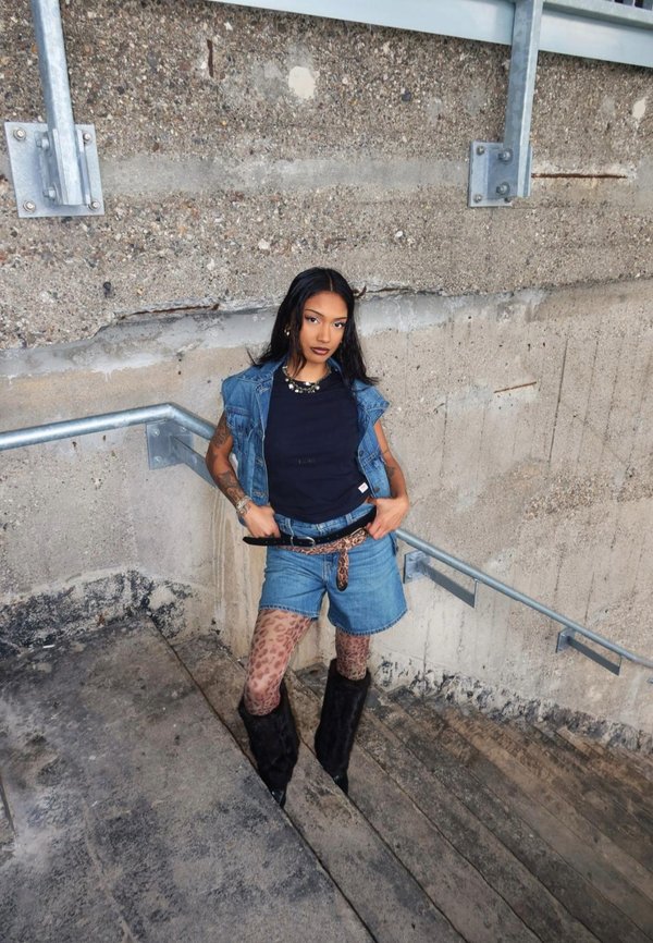 Gilet di jeans sopra una maglietta nera, abbinato a pantaloni shorts in denim a vita alta. Gli accessori includono una cintura con motivo e stivali neri di pelliccia fino al ginocchio.