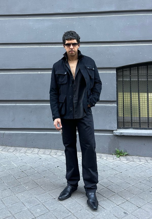 Muškarac u crnoj jakni, raskopčanoj crnoj košulji, crnim pantalonama i sunčanim naočalama stoji na sivom pločniku blizu sive zidne ograde s prozorom s rešetkama.