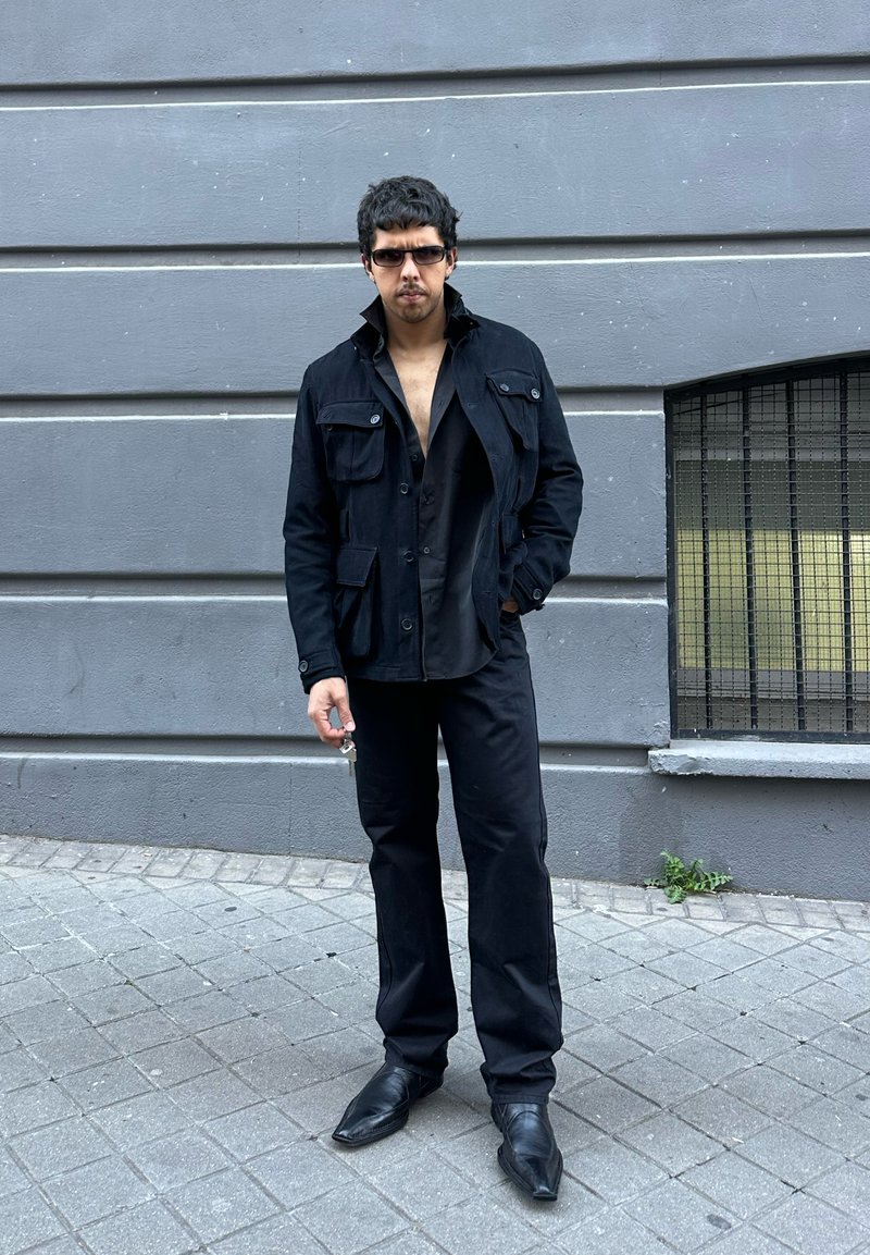 Άνδρας που φορά μαύρο μπουφάν, ανοιχτό μαύρο πουκάμισο, μαύρο παντελόνι και γυαλιά ηλίου στέκεται σε γκρι πεζοδρόμιο κοντά σε γκρι τοίχο με παράθυρο με κάγκελα.