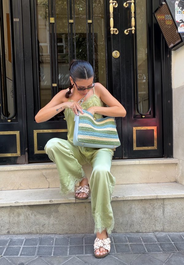 Femme en tenue satinée verte assise sur des marches, regardant dans un sac tissé rayé, portant des lunettes de soleil et des sandales avec des détails floraux.