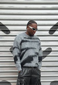 Suéter de punto gris difuso con patrones abstractos negros, combinado con pantalones de cuero negros. Gafas de sol blancas y peinado con trenzas completan el look.