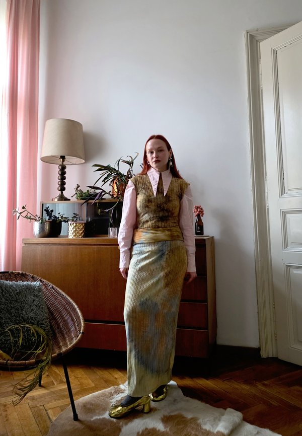 Kobieta z długimi rudymi włosami stoi w pokoju, nosząc spódnicę tie-dye, teksturowaną kamizelkę, różową bluzkę i złote buty obok drewnianej komody i roślin.