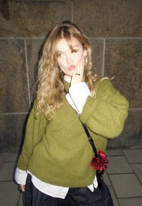 Jeune femme aux cheveux blonds ondulés portant un pull vert et une chemise blanche, tenant son visage avec la main, debout devant un mur de pierre.