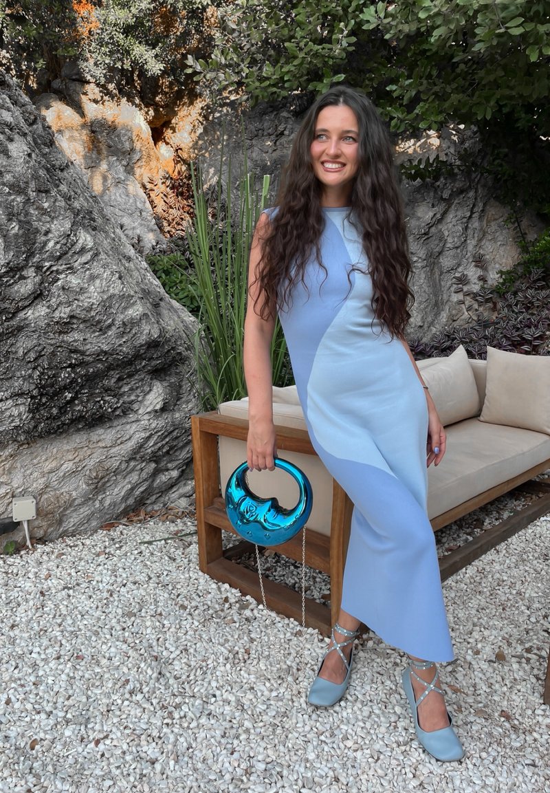 Himmelblaues, ärmelloses Kleid mit geometrischem Farbblockdesign. Als Zubehör eine türkisfarbene Handtasche mit einem runden Griff. Quadratisch geschnittene Absatzschuhe.