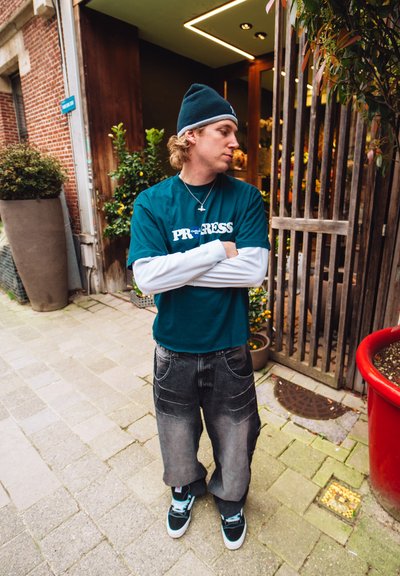 Camiseta de algodón color teal con texto blanco, superpuesta sobre una camisa de manga larga blanca, combinada con jeans holgados grises y zapatillas negras con acentos en teal.