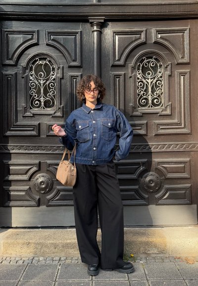 Chaqueta de mezclilla azul con cierres de botón y dos bolsillos en el pecho, combinada con pantalones anchos negros y un bolso de hombro beige frente a una puerta de madera oscura.