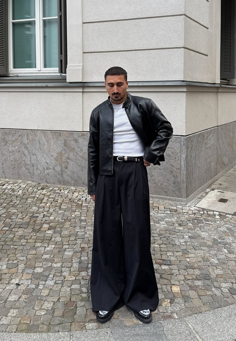 Homme portant une veste en cuir noire, une chemise blanche, un pantalon noir large et des chaussures noires, debout dans une rue pavée près d’un coin de bâtiment.