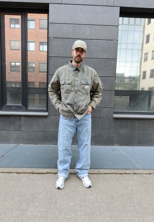 Man met een groene jas, lichtblauwe spijkerbroek, witte sneakers en een groene pet, staand op een stoep met stedelijke gebouwen op de achtergrond.