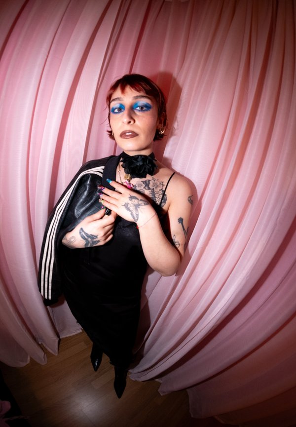 Persona con capelli corti e audace trucco occhi blu, indossando un vestito nero, che tiene una giacca, in piedi davanti a tende rosa drappeggiate.