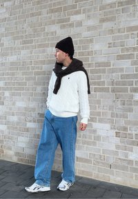 Pull en maille torsadée blanche, jeans larges bleus, bonnet noir et écharpe marron texturée. Baskets blanches et noires sur une surface pavée.