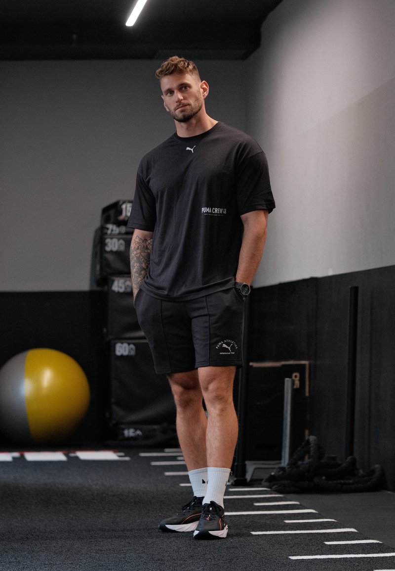T-shirt et short de sport noirs avec accents de logo. Le modèle porte des chaussures noires et des chaussettes blanches. Fond de salle de sport avec équipement d'entraînement visible.