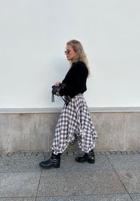 Crna pulover, suknja s smeškom u smešnim smeškom smešne ukrugle u obliku smeha, crne gležnjače s kopčama. Model stoji naslonjen na prazni zid.