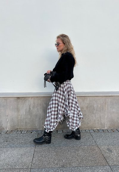Suéter negro, falda de cuadros marrón y blanca con dobladillo con volantes, botas negras de tobillo con detalles de hebilla. La modelo está de pie contra una pared en blanco.