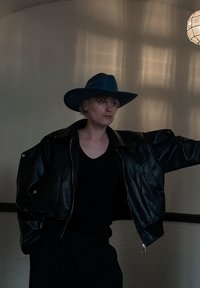 Κομμένο μαύρο δερμάτινο μπουφάν με υπερμεγέθεις ώμους, φορεμένο πάνω από μια μαύρη μπλούζα. Μπλε καπέλο με φαρδό γείσο και υφή, με φυσικά μαλλιά ορατά.